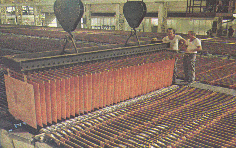 Workers stand beside a crane lifting a dense rack of copper cathode starter sheets inside a refinery.