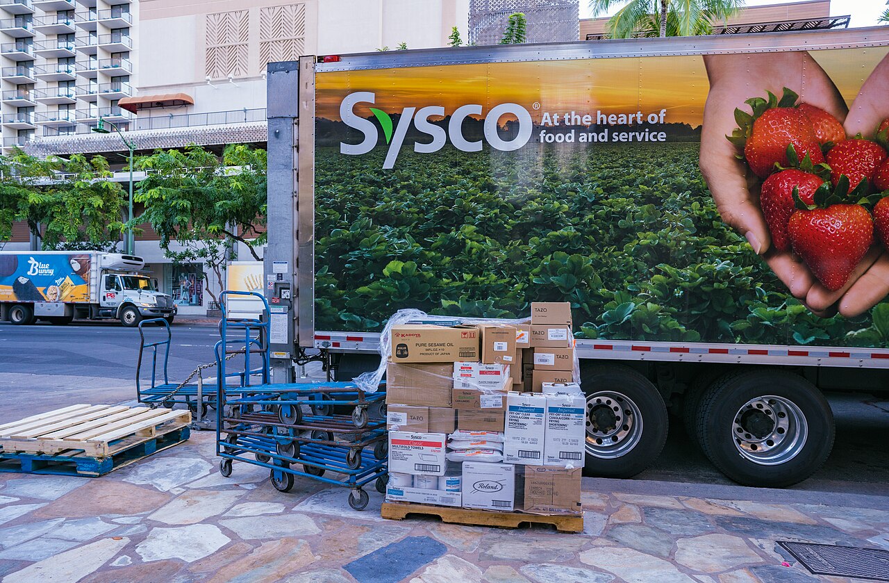 A Sysco foodservice delivery truck with a liftgate and pallet being unloaded beside a curb.
