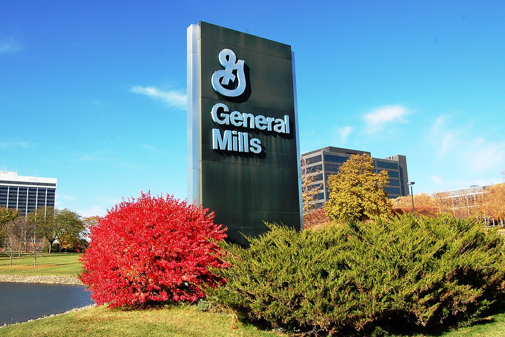 阳光下，明尼苏达州 Golden Valley 的 General Mills 总部外立着公司标牌，周围可见办公楼、灌木和池塘。