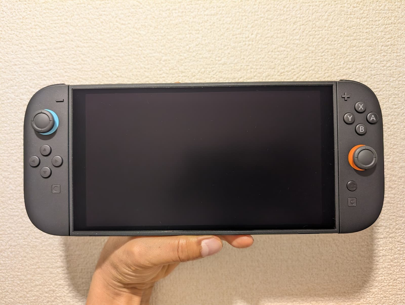 一只手横持掌机模式下的 Nintendo Switch 2，两侧摇杆周围分别带有蓝色与橙色点缀。