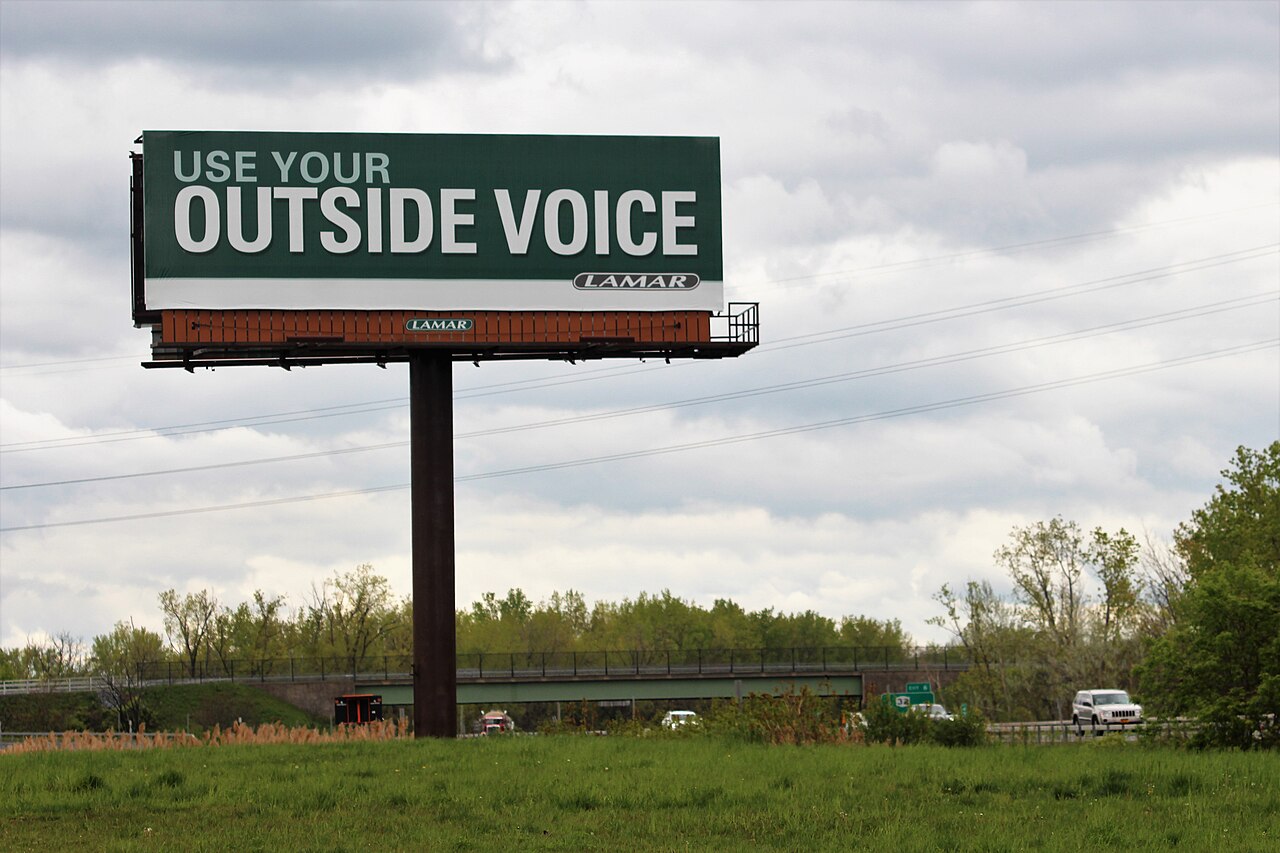 A Lamar roadside billboard in Menands, New York, standing over grass, trees, traffic, and a cloudy sky.