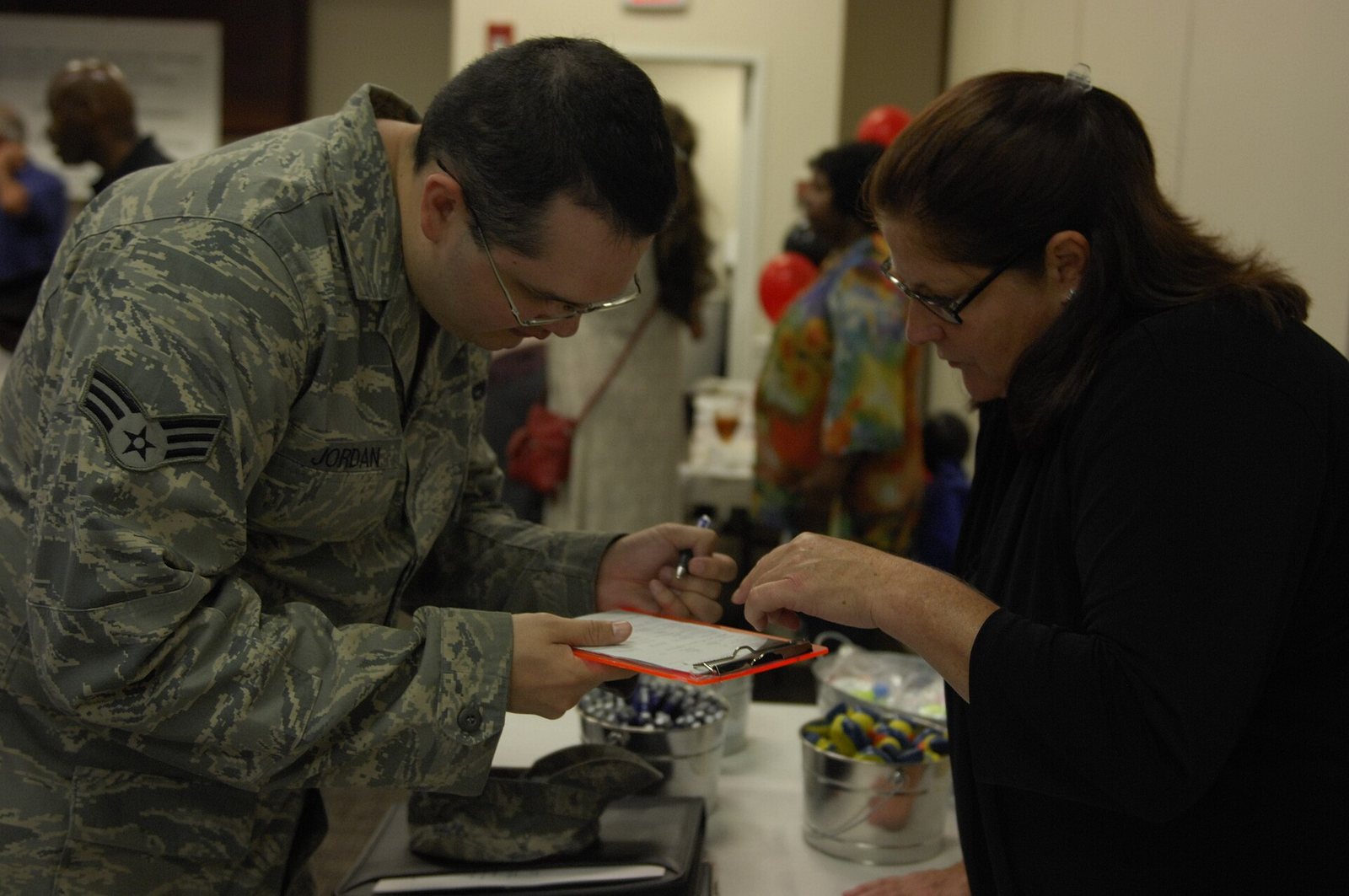 A U.S. Air Force service member and a civilian recruiter lean over a clipboard at a busy indoor job fair.