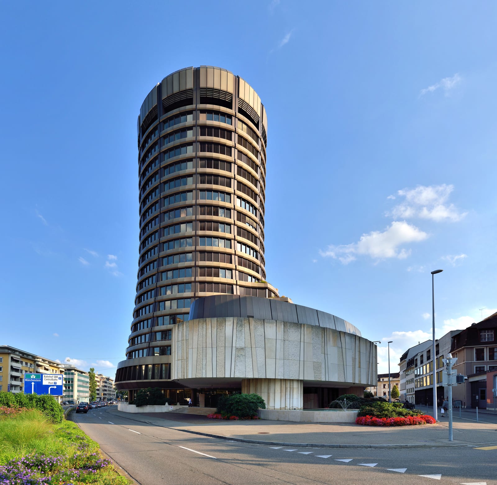 The cylindrical Bank for International Settlements tower rising above a Basel street under a clear blue sky.