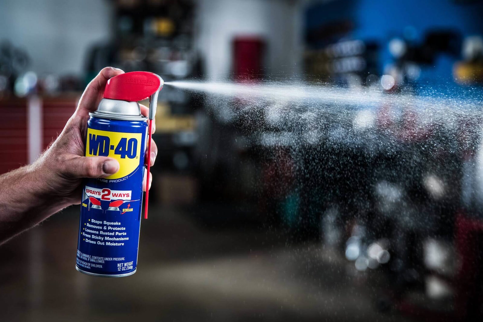 A hand sprays WD-40 from a Smart Straw aerosol can across dark workshop equipment.