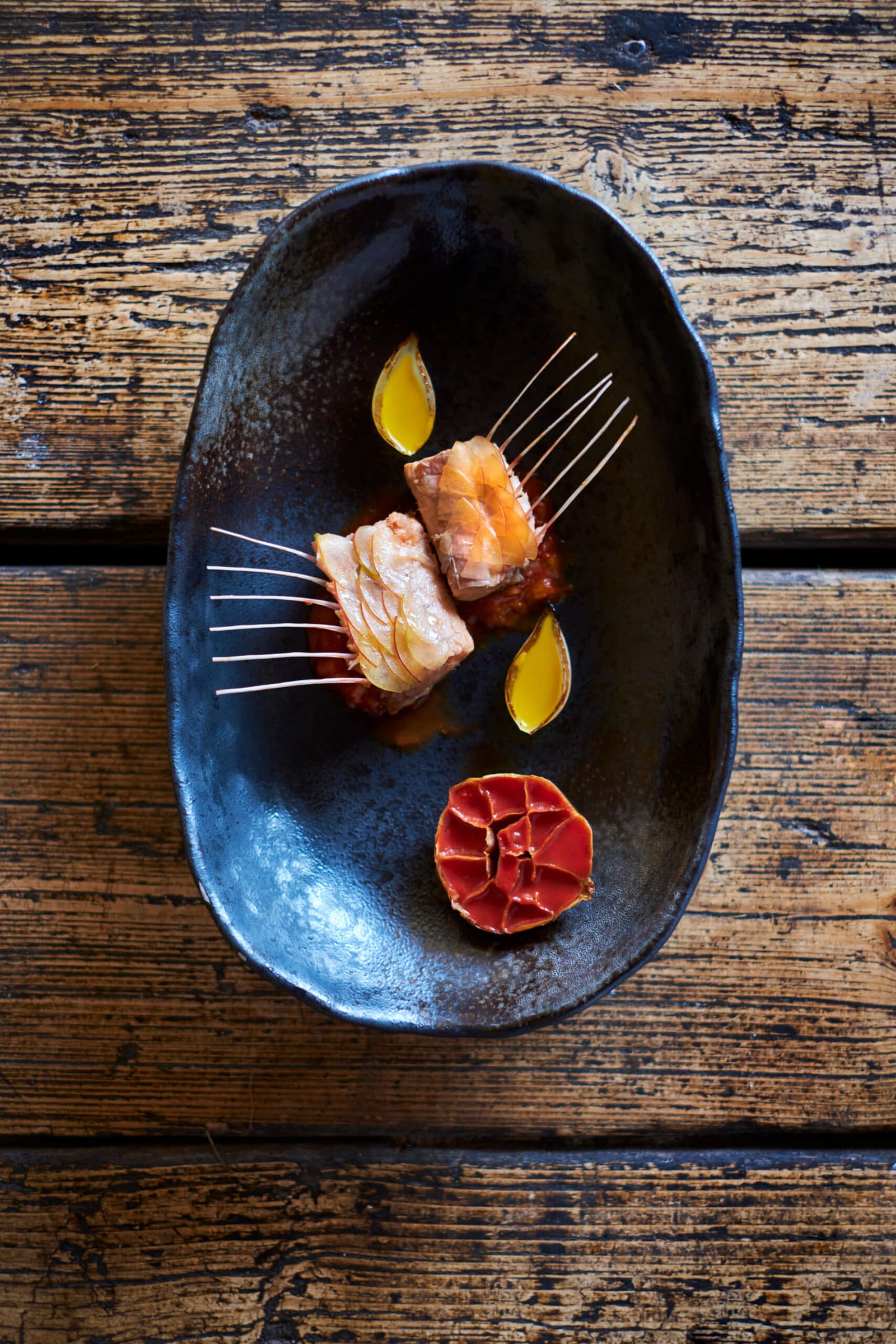 Two pieces of prepared carp in a long dark ceramic dish with amber sauce and garnishes, photographed on a weathered wooden table.
