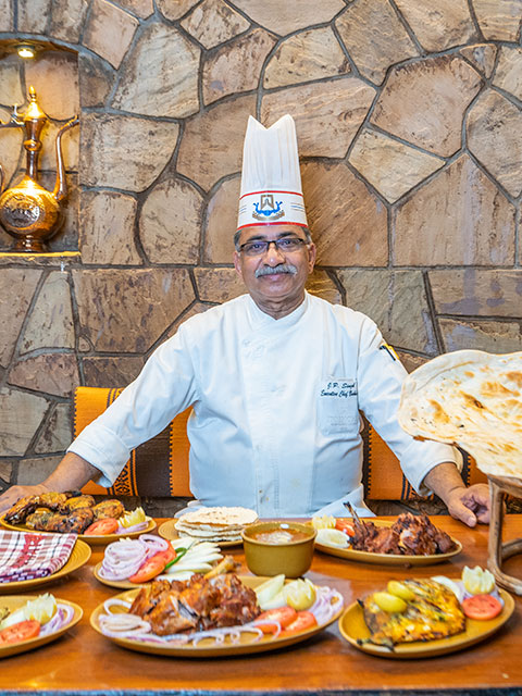 A Bukhara chef at ITC Maurya seated behind naan, dal, kebabs, and tandoori dishes in the restaurant's stone-walled dining room.