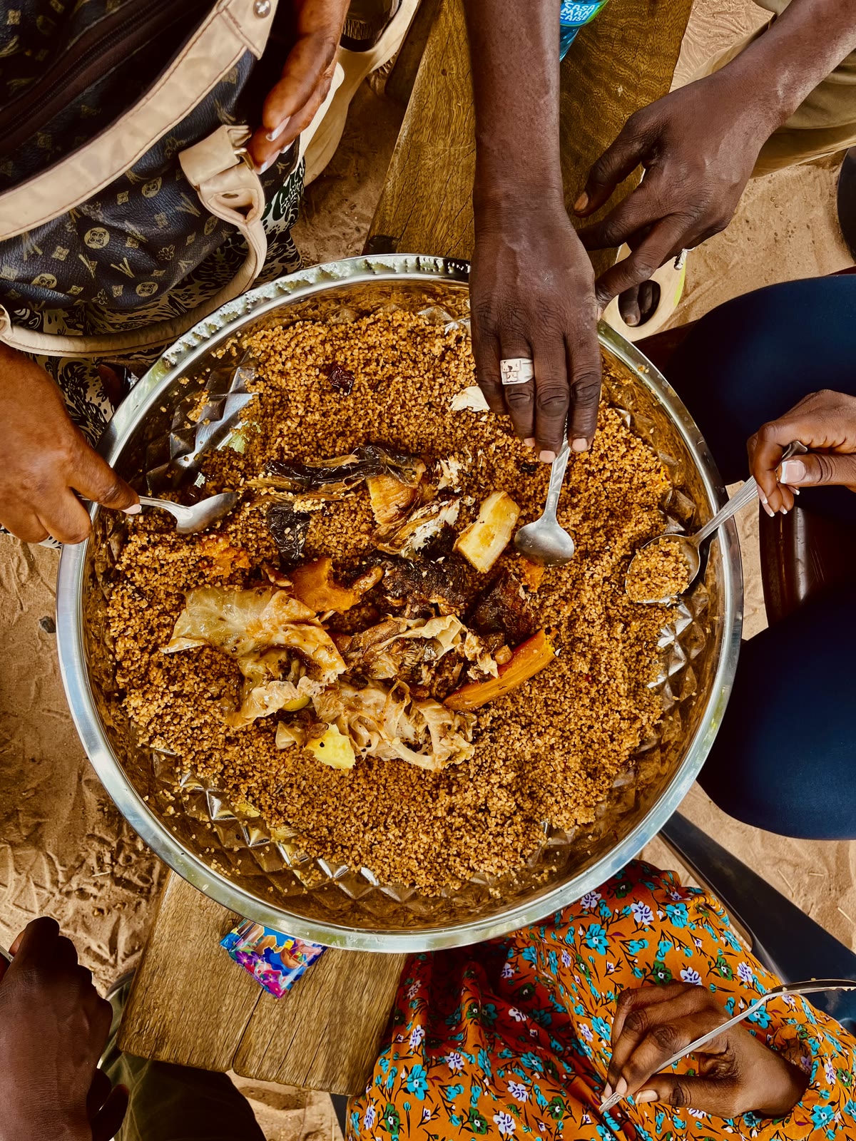 Hands holding spoons around a shared aluminum platter of red ceebu jën with fish and large pieces of vegetables.
