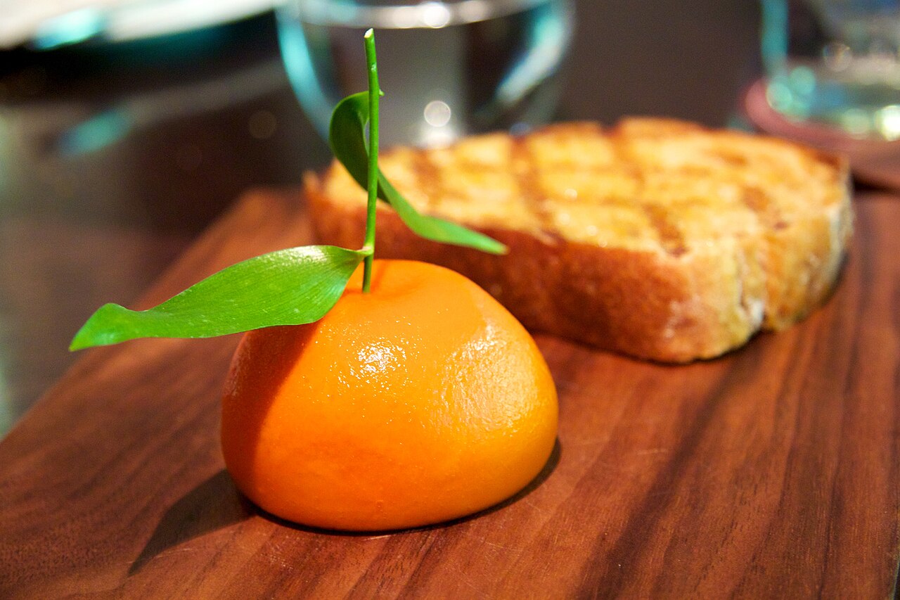 A glossy orange Meat Fruit mandarin with green leaves, served beside grilled bread on a wooden board.