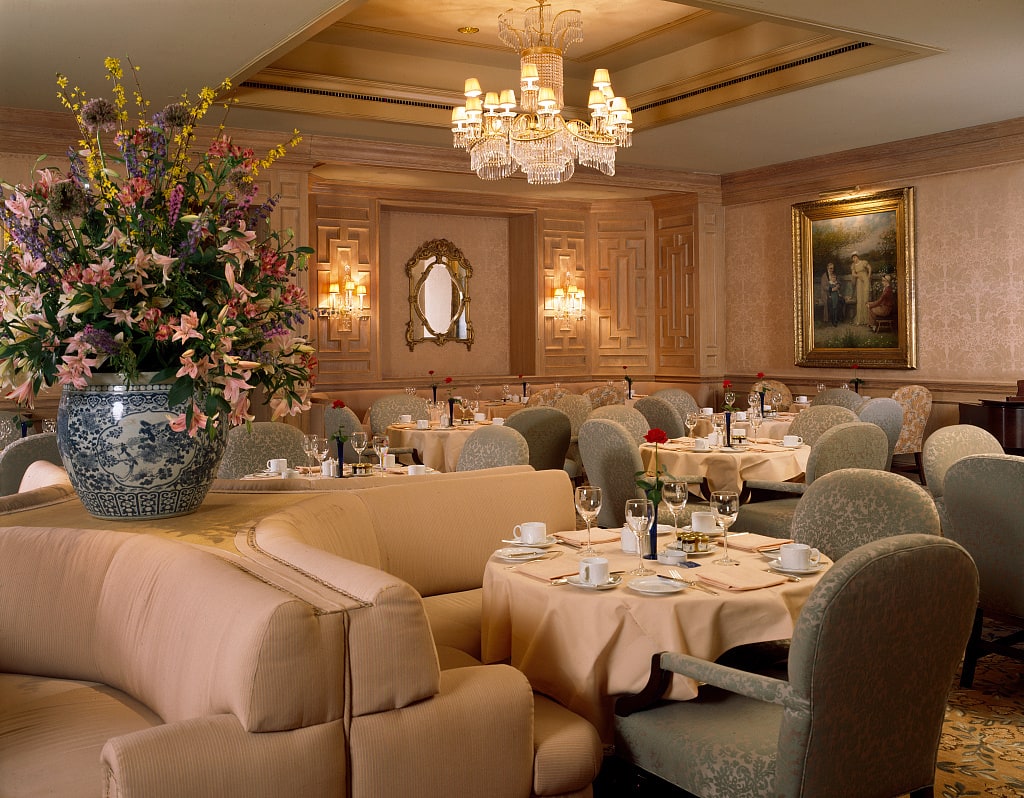 A formally set American restaurant dining room photographed in the 1980s, with white napery, flowers, crystal, upholstered chairs, and a chandelier.