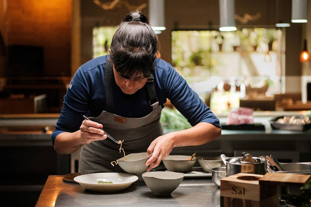 Chef Manu Buffara bends over a white bowl while plating at the kitchen pass in Curitiba.