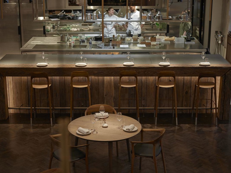 Nineteen18 dining room in Vilnius with a long counter, set tables, warm wood, and chefs visible through the open kitchen pass.