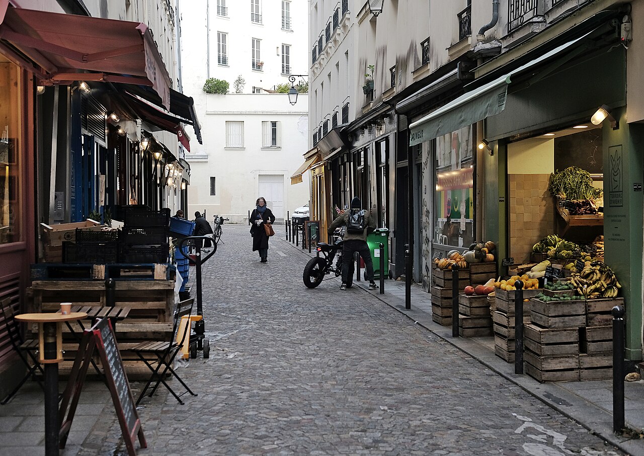 A narrow cobbled Paris street lined with food shops, with wooden produce crates outside a greengrocer and pedestrians in the lane.