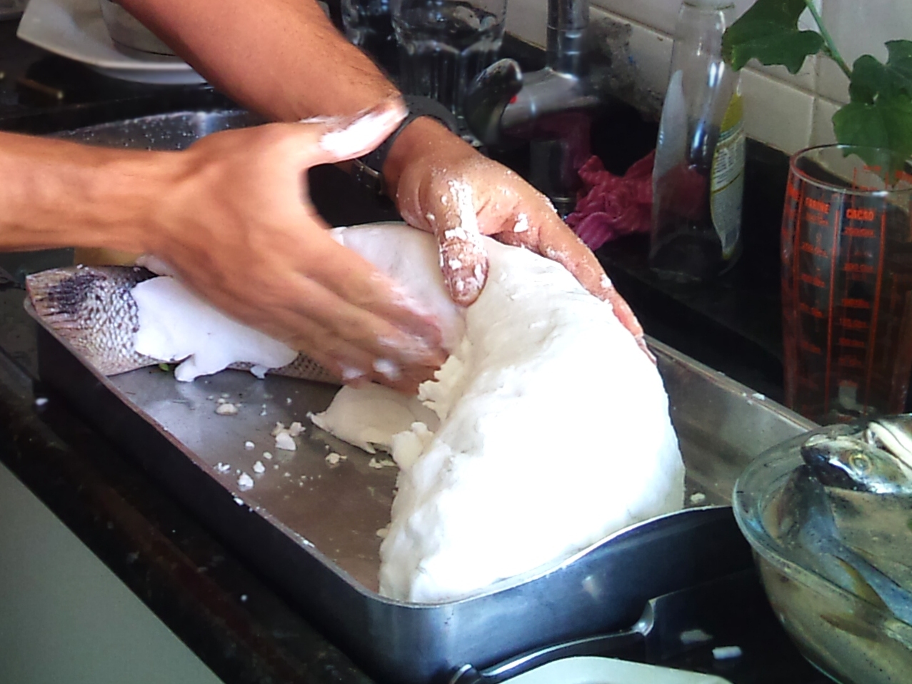 Hands press a thick pale salt mixture over a whole fish on a metal tray before baking.