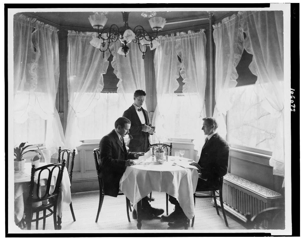 A waiter stands beside two seated diners while taking their order in a restaurant around 1906.