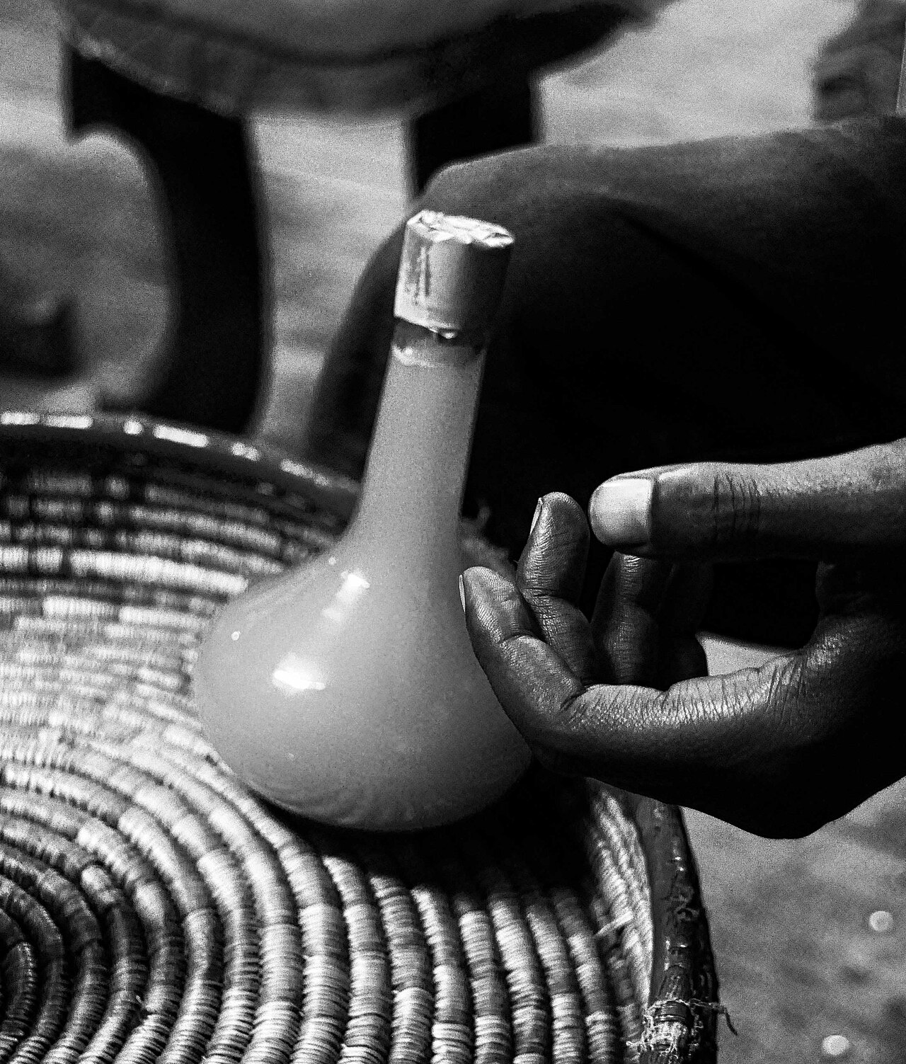 Black-and-white close-up of a hand beside a round-bellied bottle of Ethiopian tej resting in a woven basket.