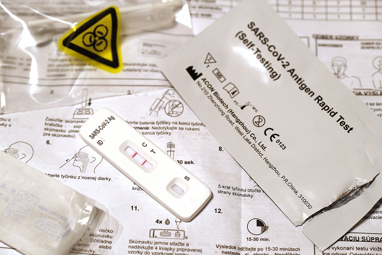 A positive COVID-19 rapid antigen test cassette on a table next to its buffer vial and packaging.