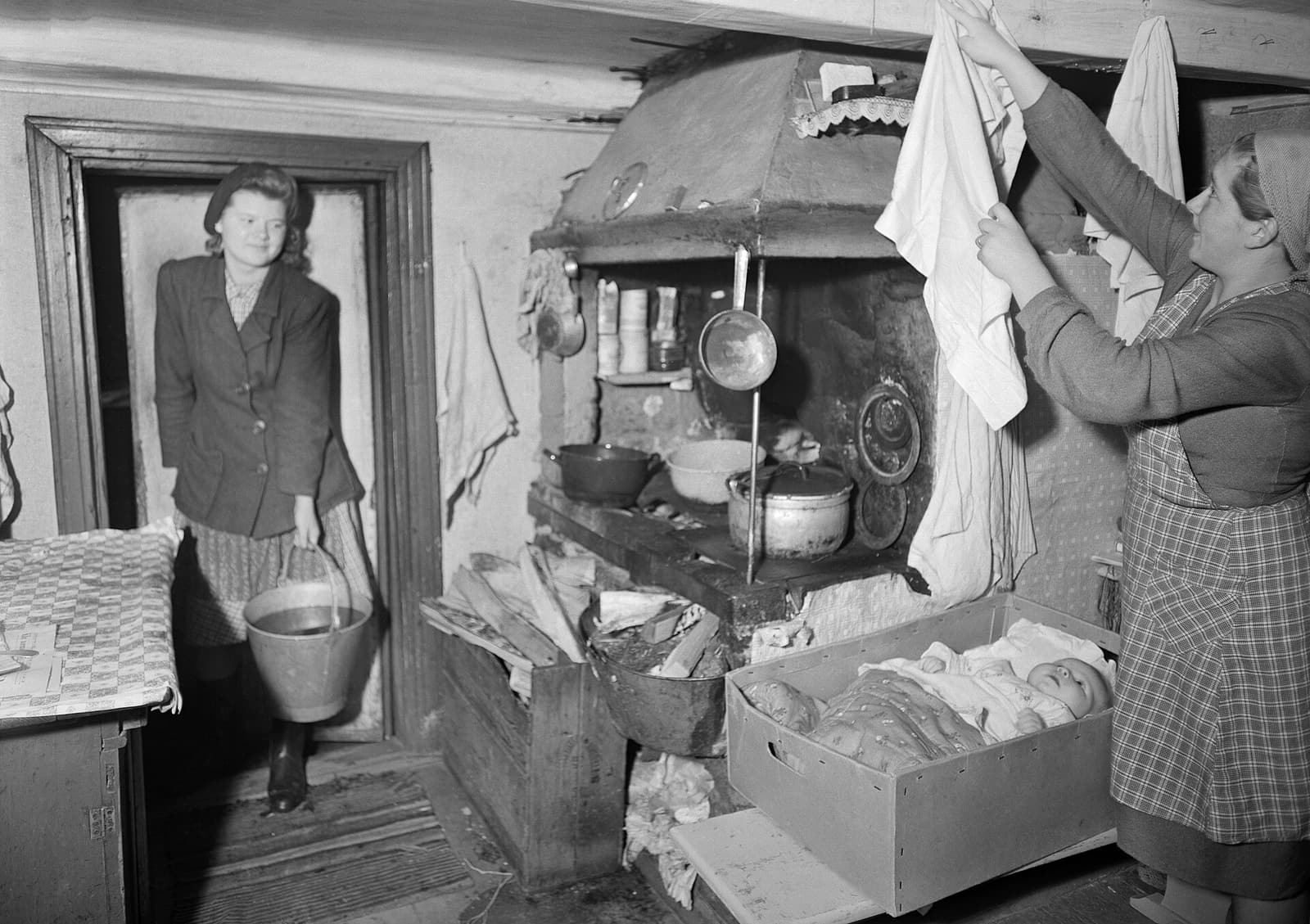 Black-and-white 1948 photograph inside a Finnish home, where a baby lies in an open maternity-package box while her mother holds up an infant garment.