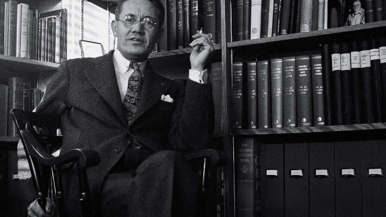 Black-and-white archival portrait of physician Henry K. Beecher seated before shelves of bound medical journals in 1950.