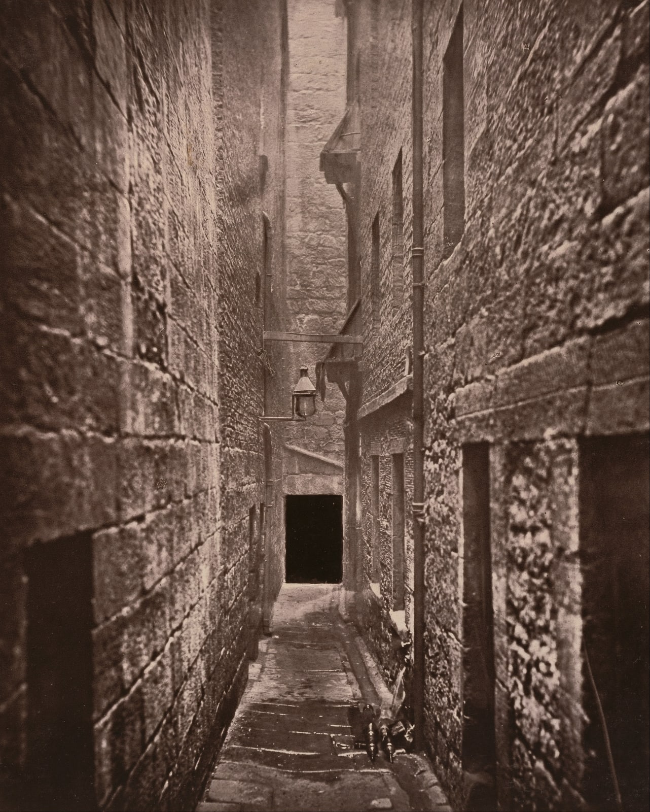 Sepia photograph looking down a narrow stone-walled Glasgow close, with a drainage channel running along the passage and little daylight reaching the ground.