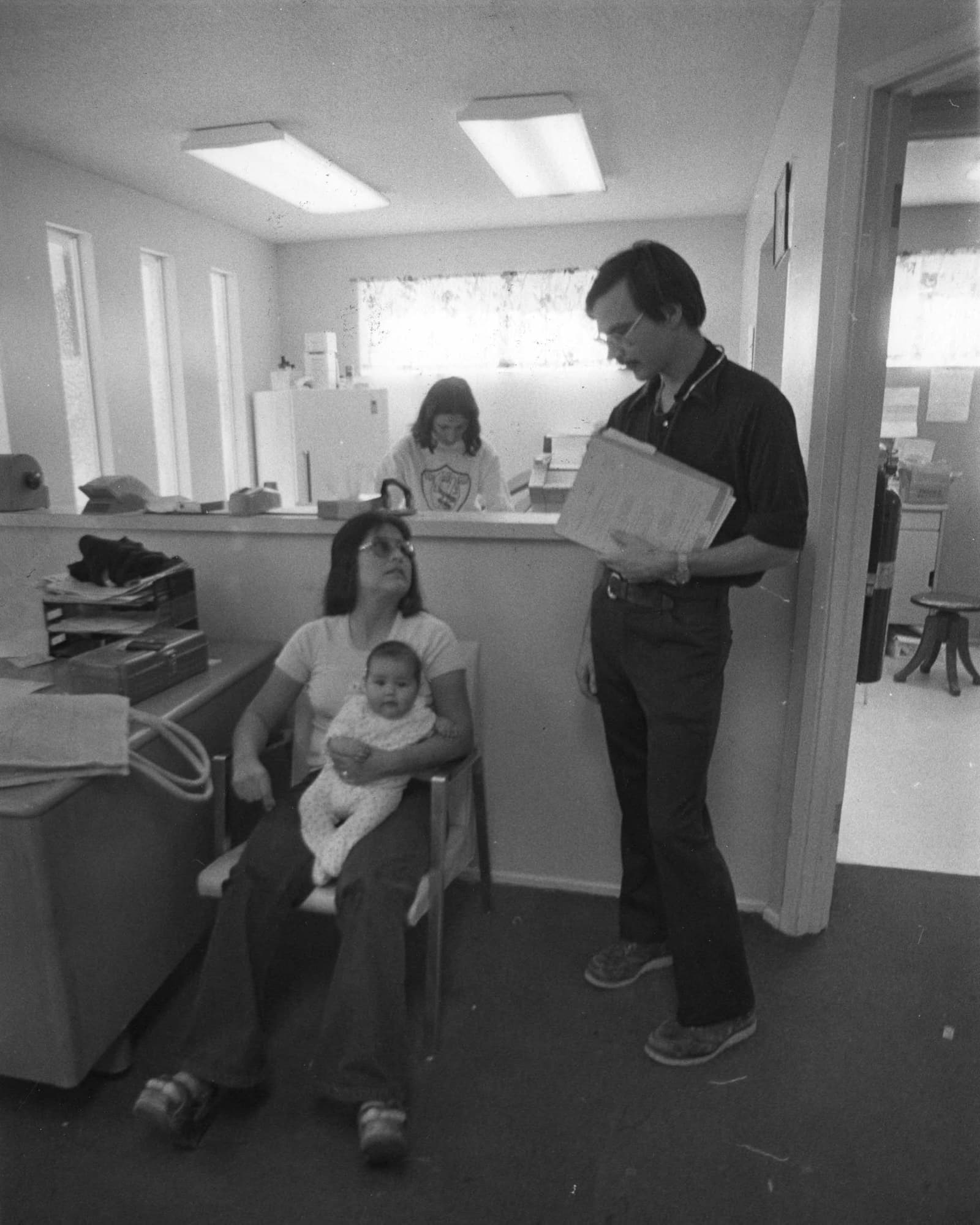 Black-and-white late-1970s photograph of a doctor holding a chart while speaking with a seated mother holding her baby at the Calexico Clinic.