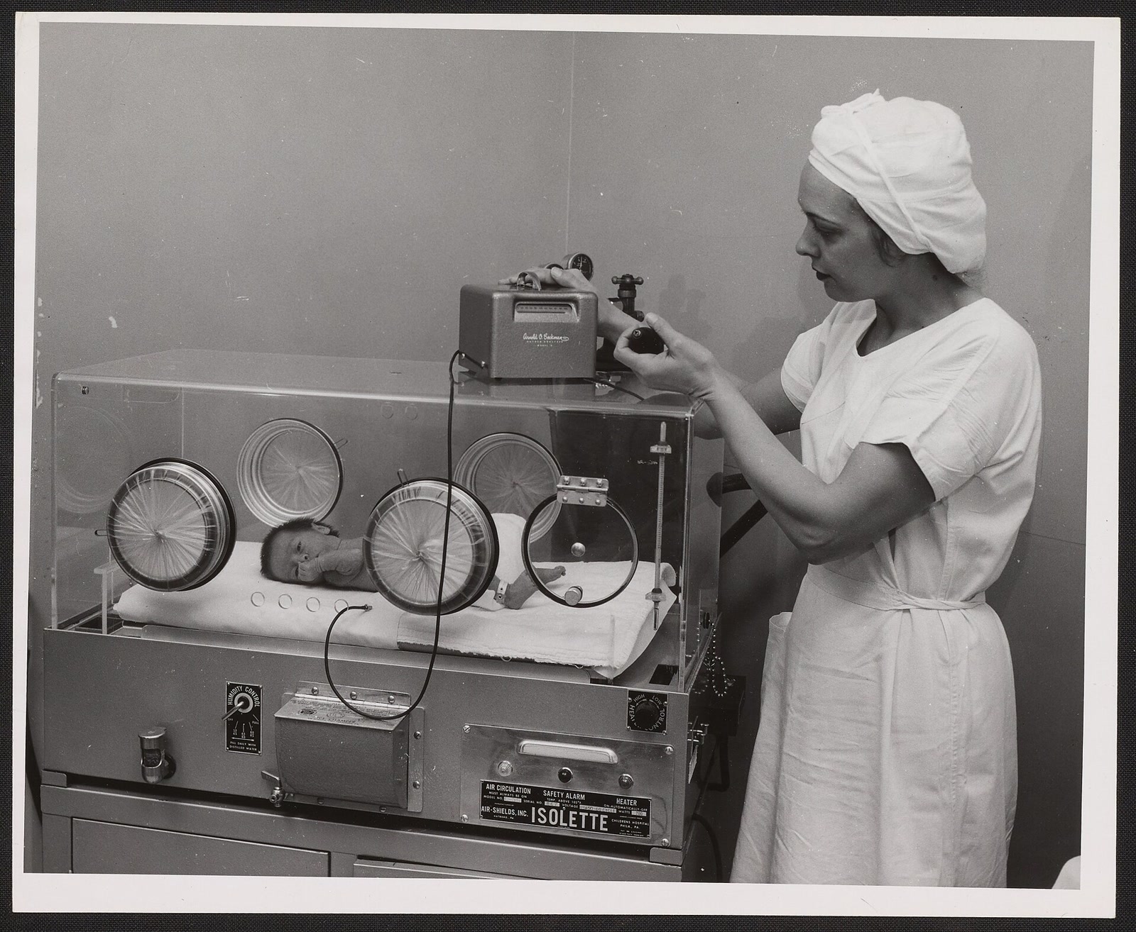 Black-and-white archival photograph of a nurse reading a Beckman oxygen meter beside a premature infant lying in a hospital incubator.