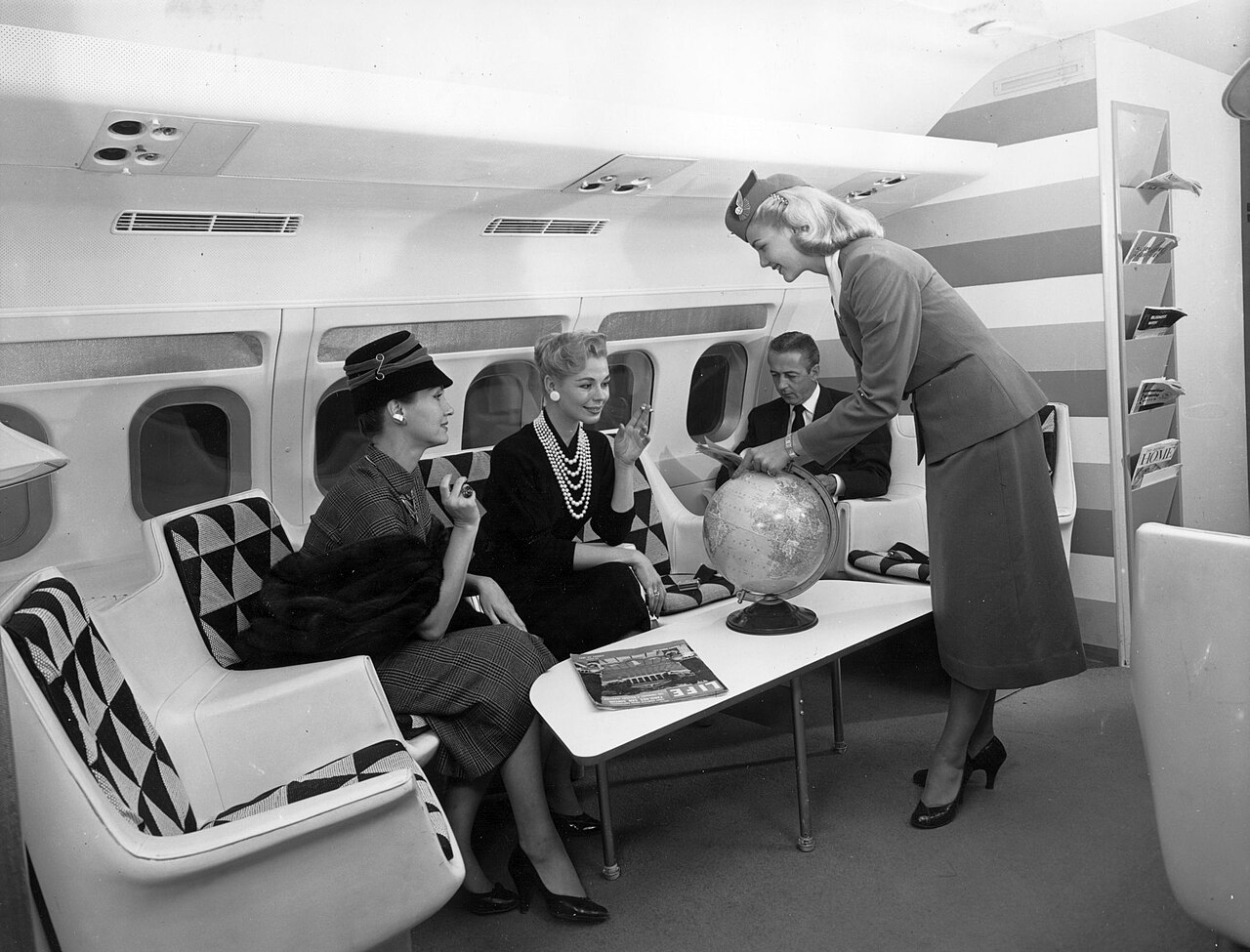 A staged 1958 TWA cabin publicity photograph showing a seated passenger holding a cigarette while a flight attendant points to a globe.
