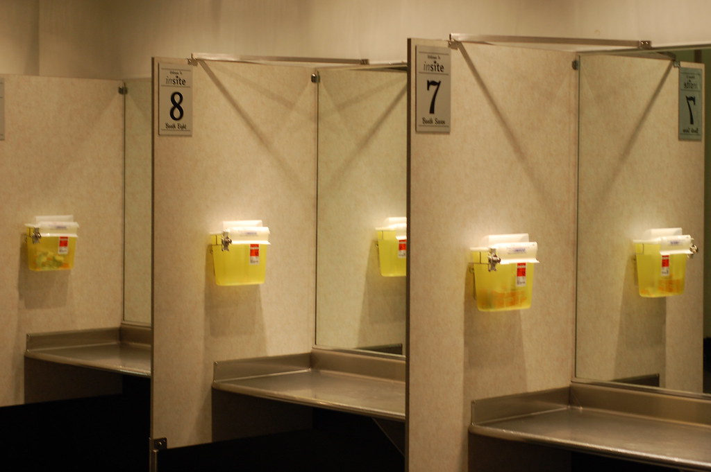 Numbered injection booths at Vancouver's Insite, separated by beige partitions and fitted with stainless-steel counters and yellow sharps bins.