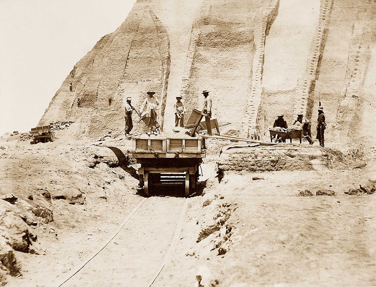 Sepia-toned archival photograph of guano miners loading a rail cart beside steep cut faces in the Great Heap on Peru's Chincha Islands.