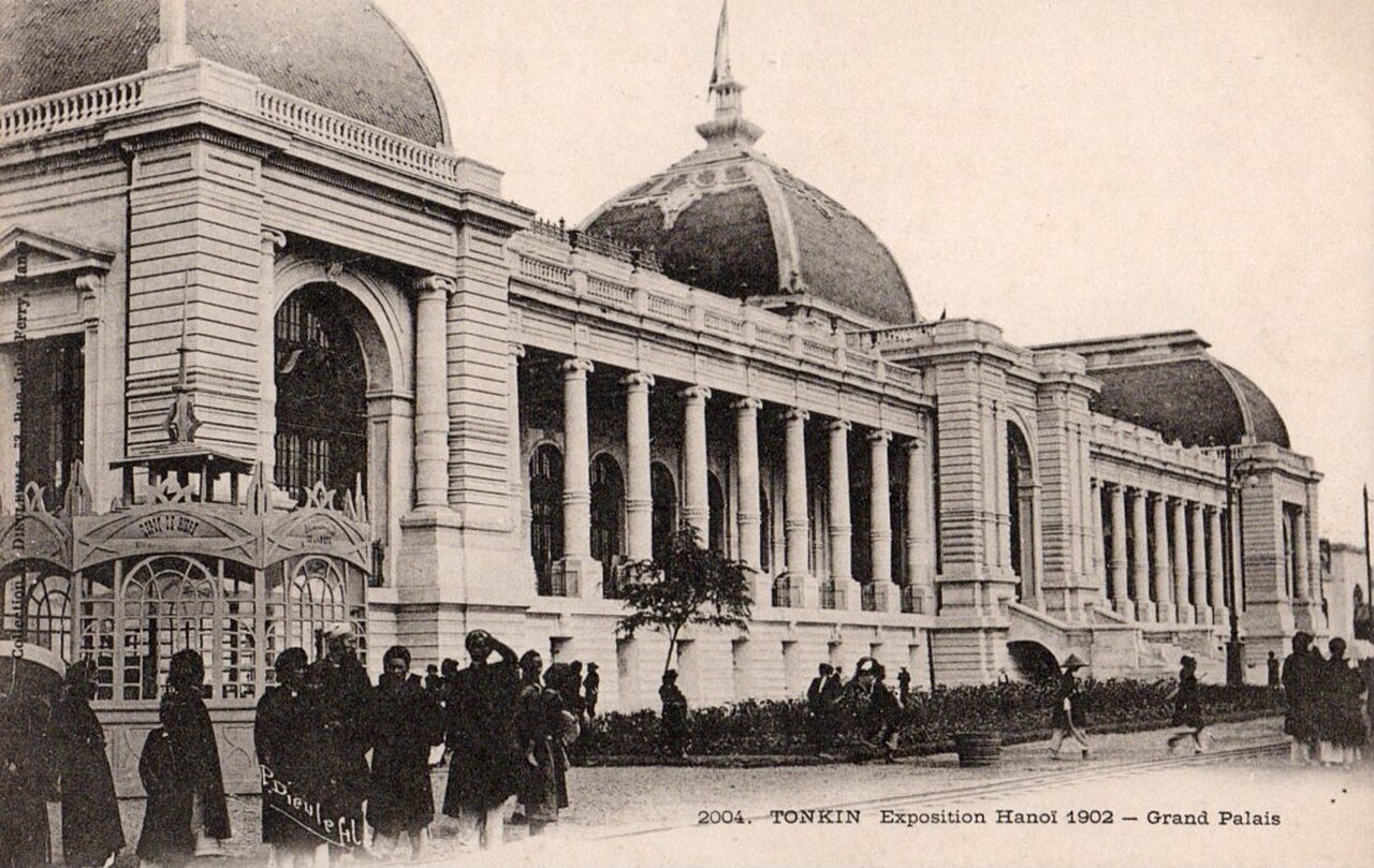 一张摄于 1902 年的黑白档案照片，画面是河内大展览宫：狭长的展馆立面、中央穹顶、规整的前庭，以及散布在建筑外的几小群人。