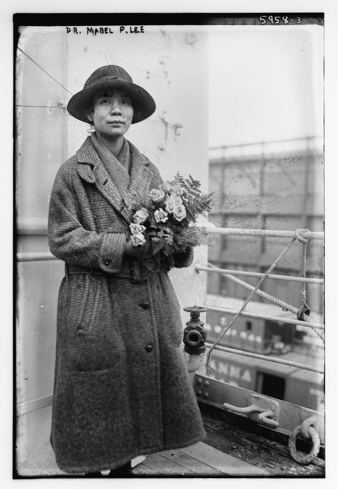 Black-and-white photograph of Mabel Ping-Hua Lee in a hat and overcoat, holding a bouquet on an outdoor deck.