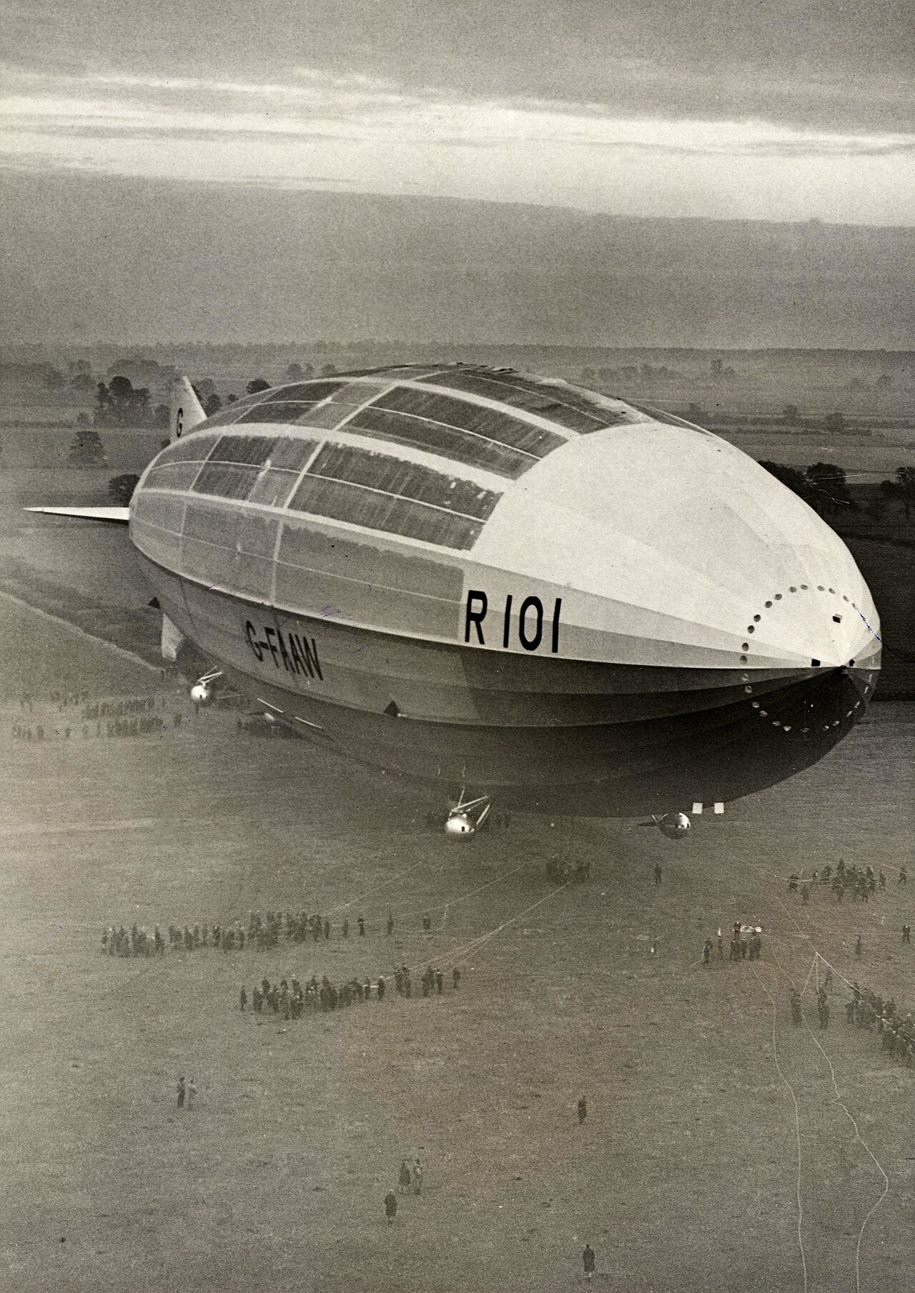 Black-and-white archival photograph looking down across the rounded nose and immense hull of R101, with groups of people appearing tiny on the ground below.