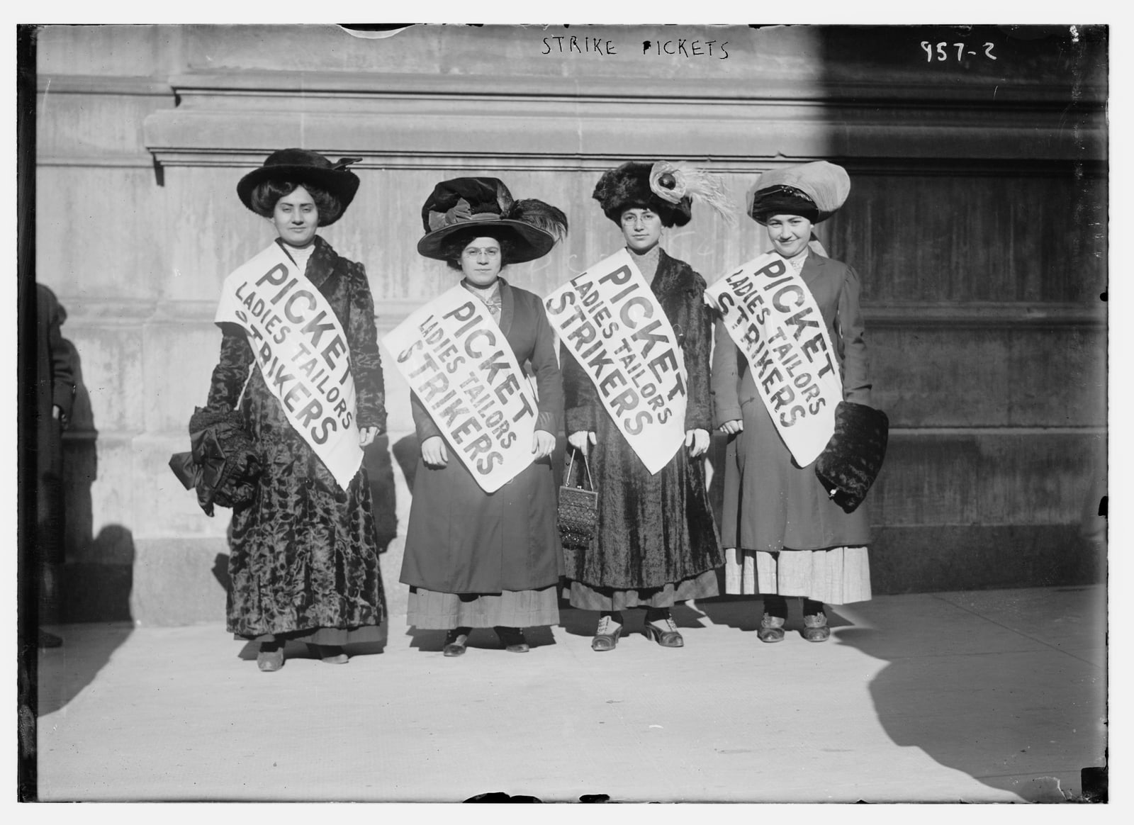 Four women garment strikers wearing diagonal picket sashes stand in winter sunlight outside a stone building in New York in February 1910.