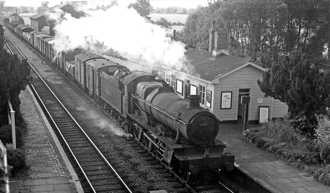 Black-and-white 1961 photograph of Adlestrop station, with a steam freight train beside the platform and station buildings beyond.