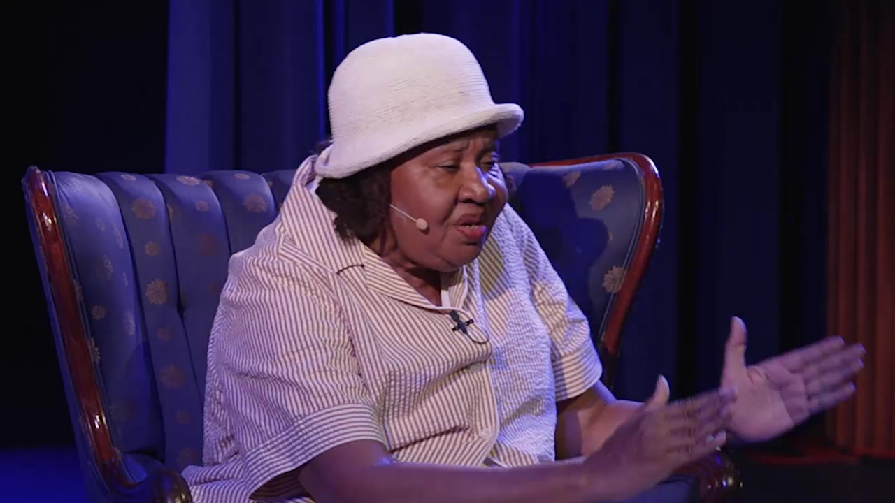 Jamaica Kincaid gestures with both hands while speaking onstage in a blue armchair.