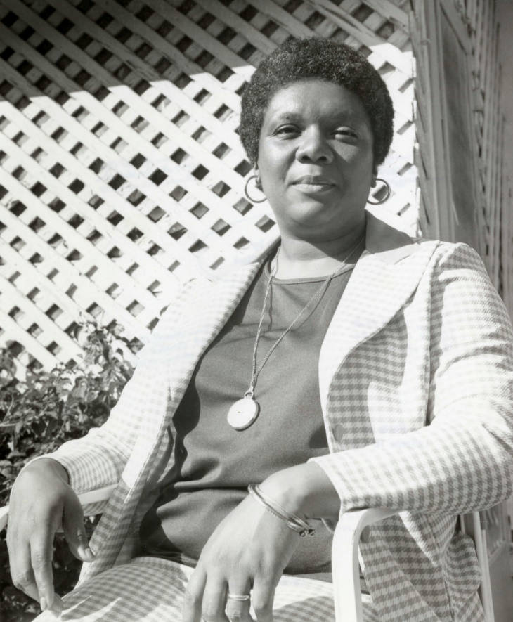 Black-and-white 1975 portrait of Lucille Clifton seated outdoors in a checkered jacket before a white lattice.