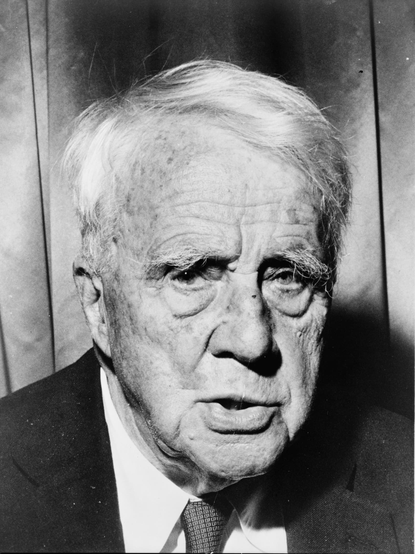 Black-and-white press photograph of Robert Frost in later life, wearing a suit and looking to the side.