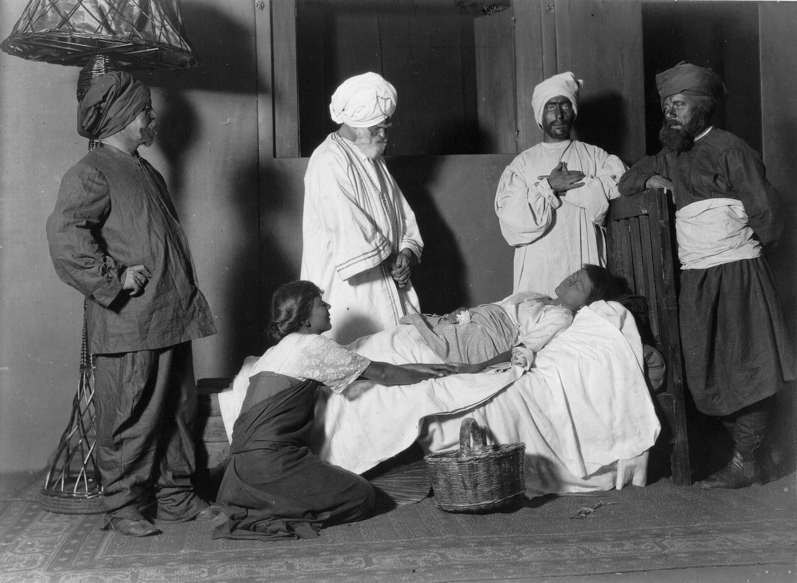 Black-and-white 1915 stage photograph of six actors gathered around Amal's bed in a Toronto production of Rabindranath Tagore's The Post Office; several performers wear turbans and dark makeup.