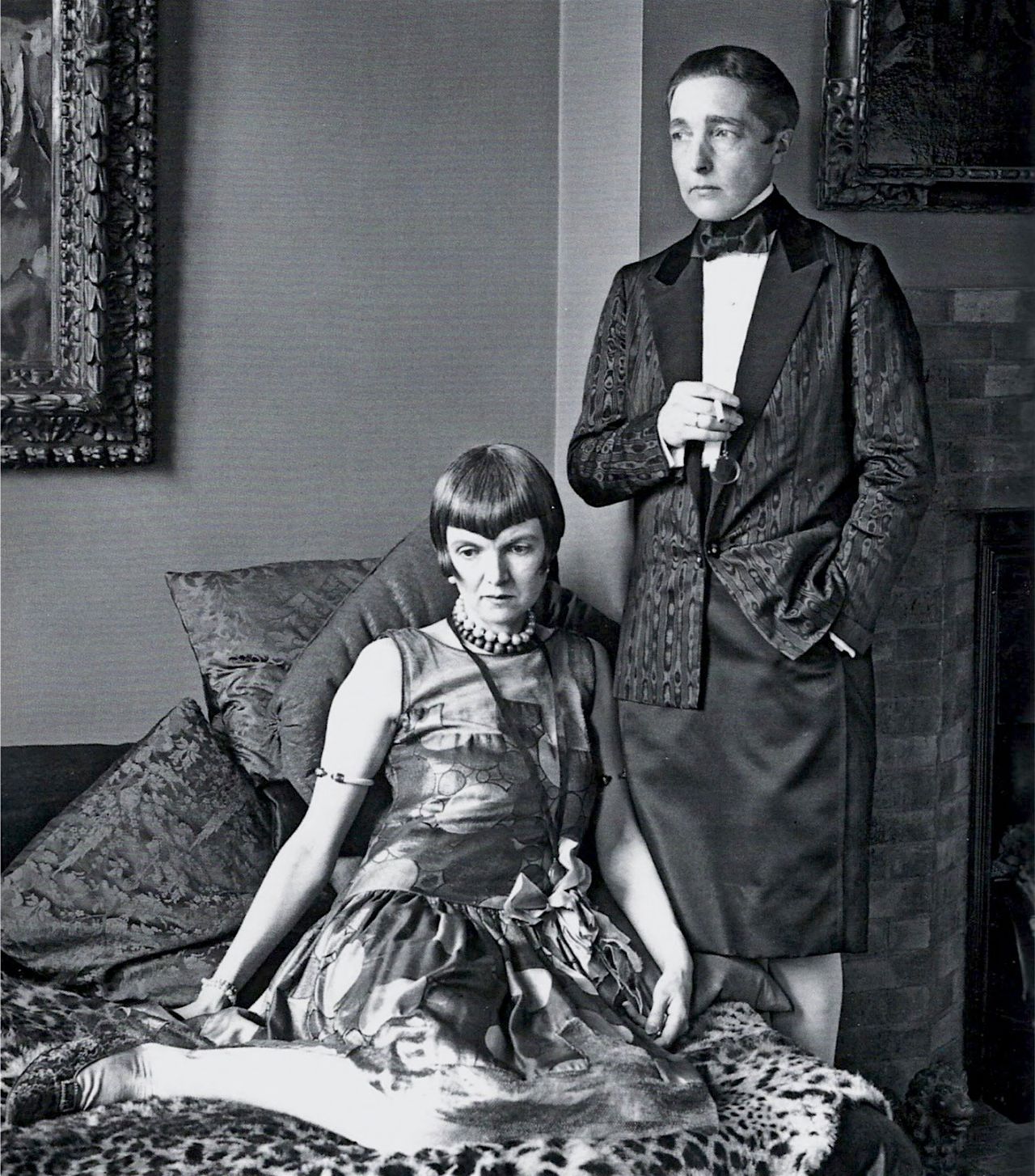 Black-and-white archival photograph of Radclyffe Hall standing beside Una Troubridge, who reclines in formal dress, in 1927.