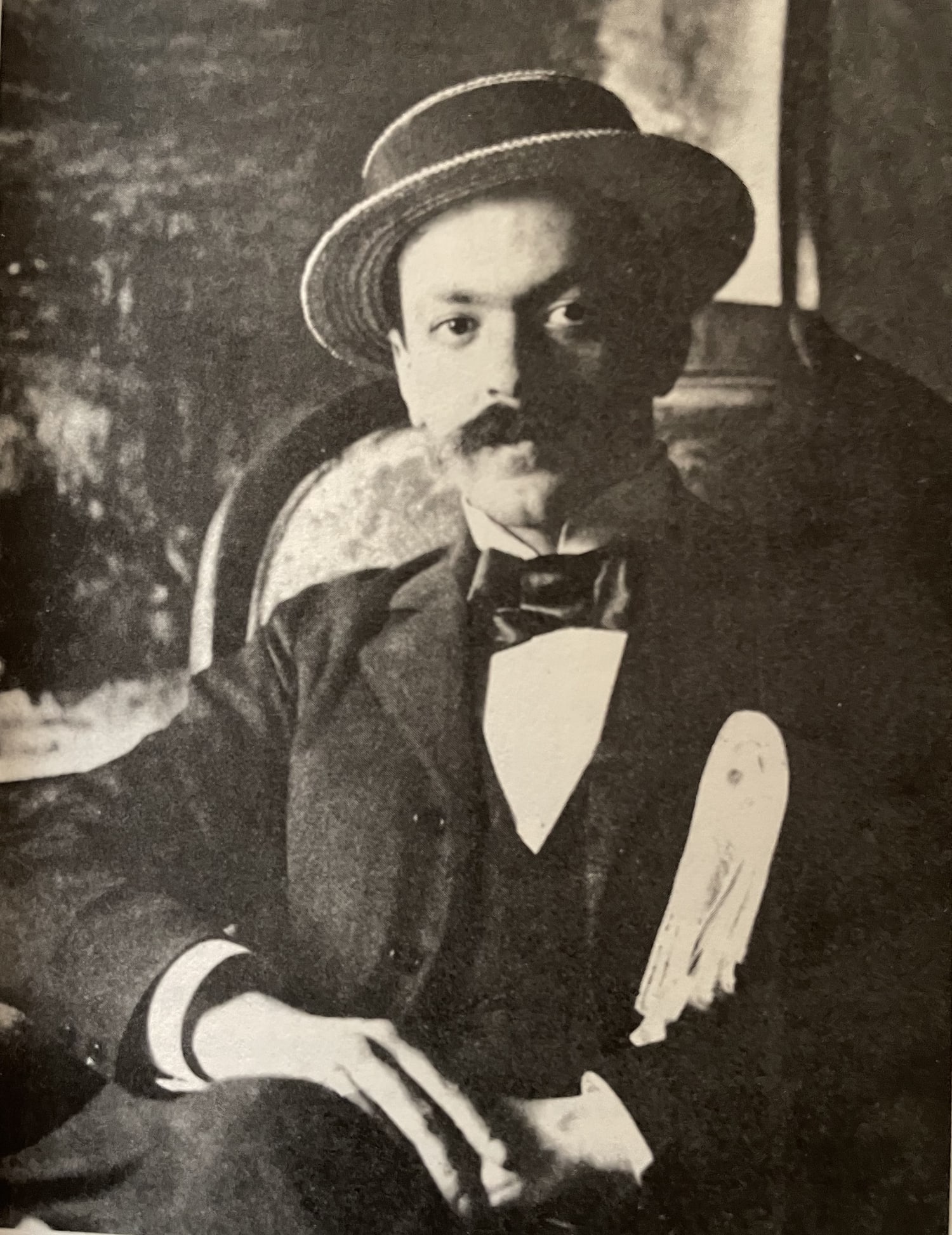 Sepia-toned archival portrait of Italo Svevo seated in a dark suit and bow tie beneath a wide-brimmed straw hat around 1920.
