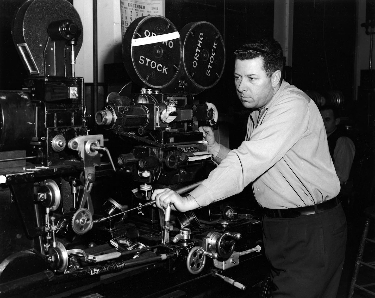 Linwood G. Dunn sits at an optical printer in a 1940s film laboratory, surrounded by reels, sprockets, lenses, and mechanical controls.