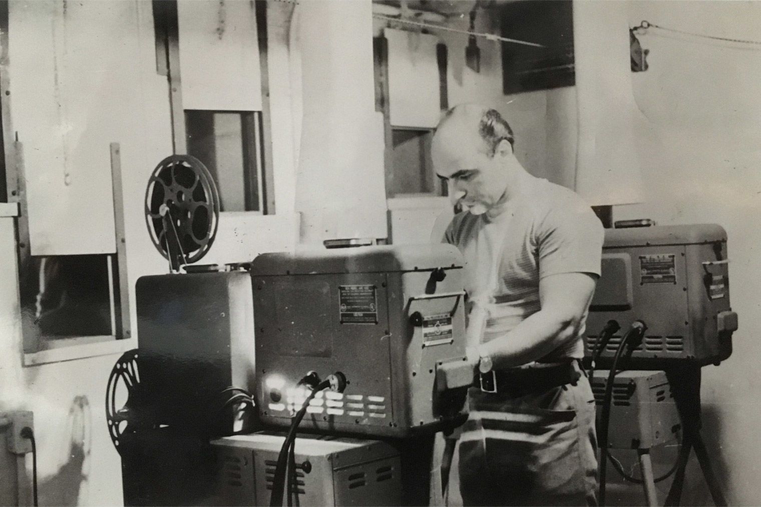 Black-and-white archival photograph of artist and audio-visual technician Ray Cusie trimming the carbon arc of an RCA Porto-Arc 400 16-millimeter projector in the Metropolitan Museum of Art's Junior Museum auditorium booth.