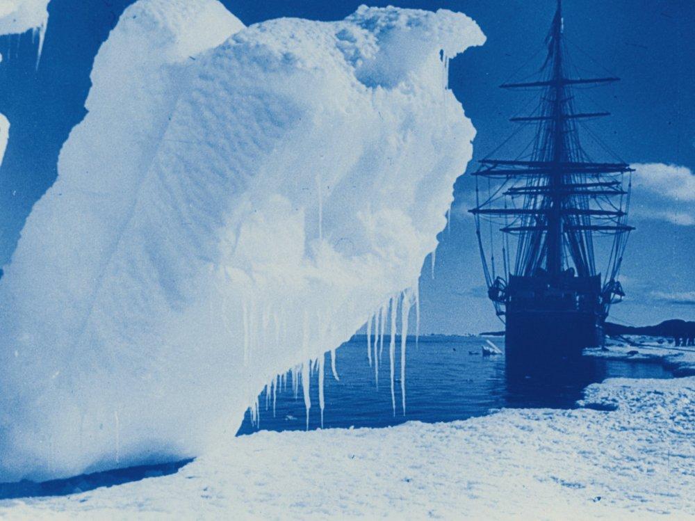 A blue-tinted archival frame from The Great White Silence, with a massive ridged iceberg filling the foreground and the Terra Nova beyond it.