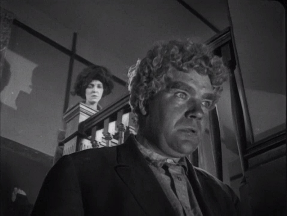 Black-and-white frame from Greed showing Gibson Gowland as McTeague in the foreground while ZaSu Pitts as Trina stands behind him on a stair landing.
