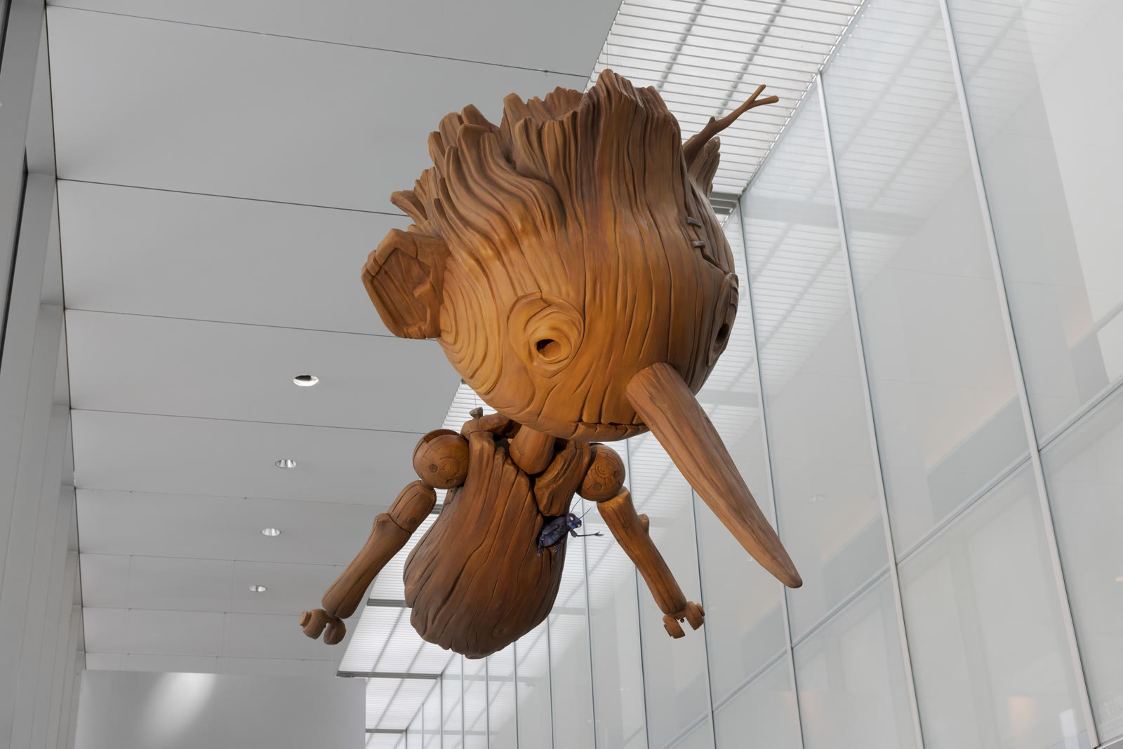 A museum installation photograph looking up at an enlarged wooden Pinocchio figure with a small blue Sebastian J. Cricket perched on his chest at MoMA.