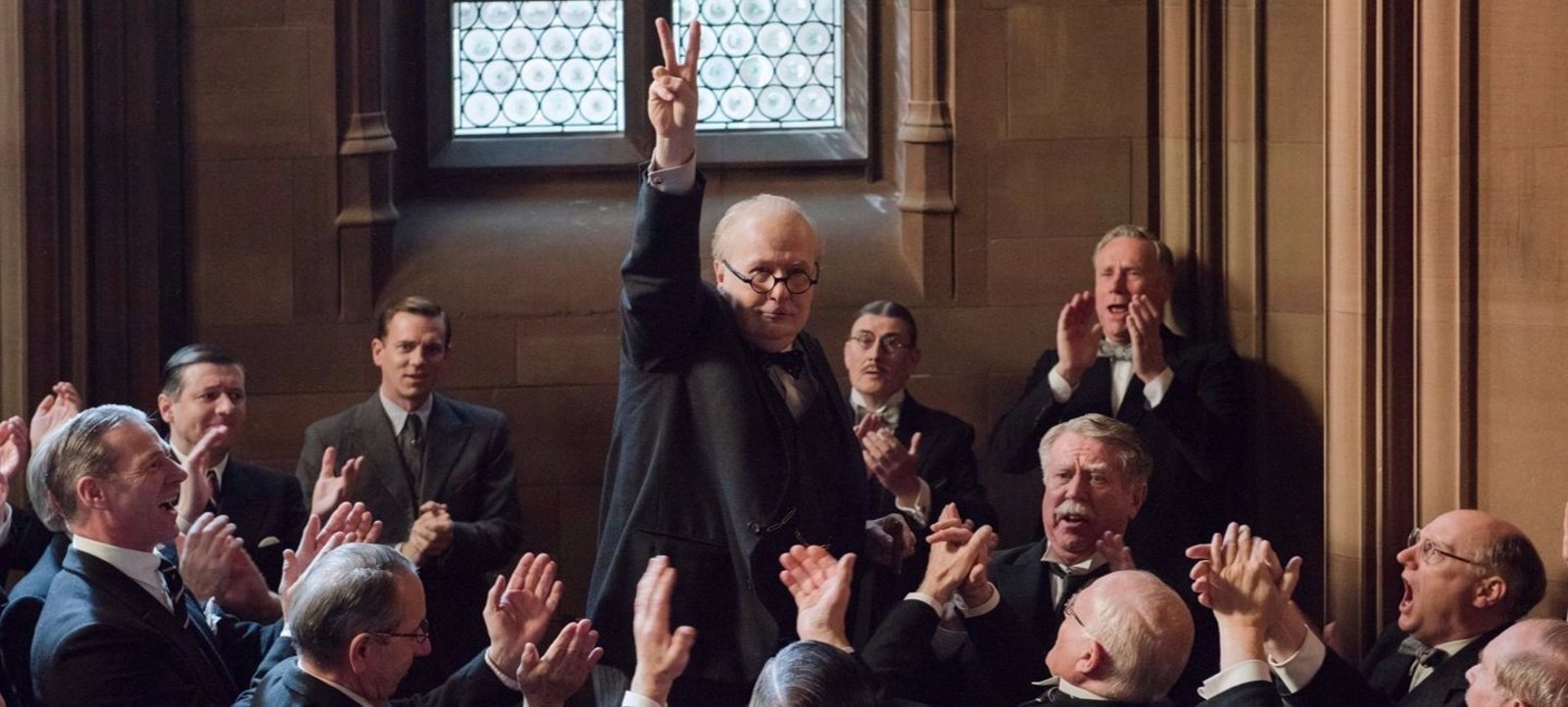 Gary Oldman as Winston Churchill raises a V sign while men around him applaud in a scene from Darkest Hour.