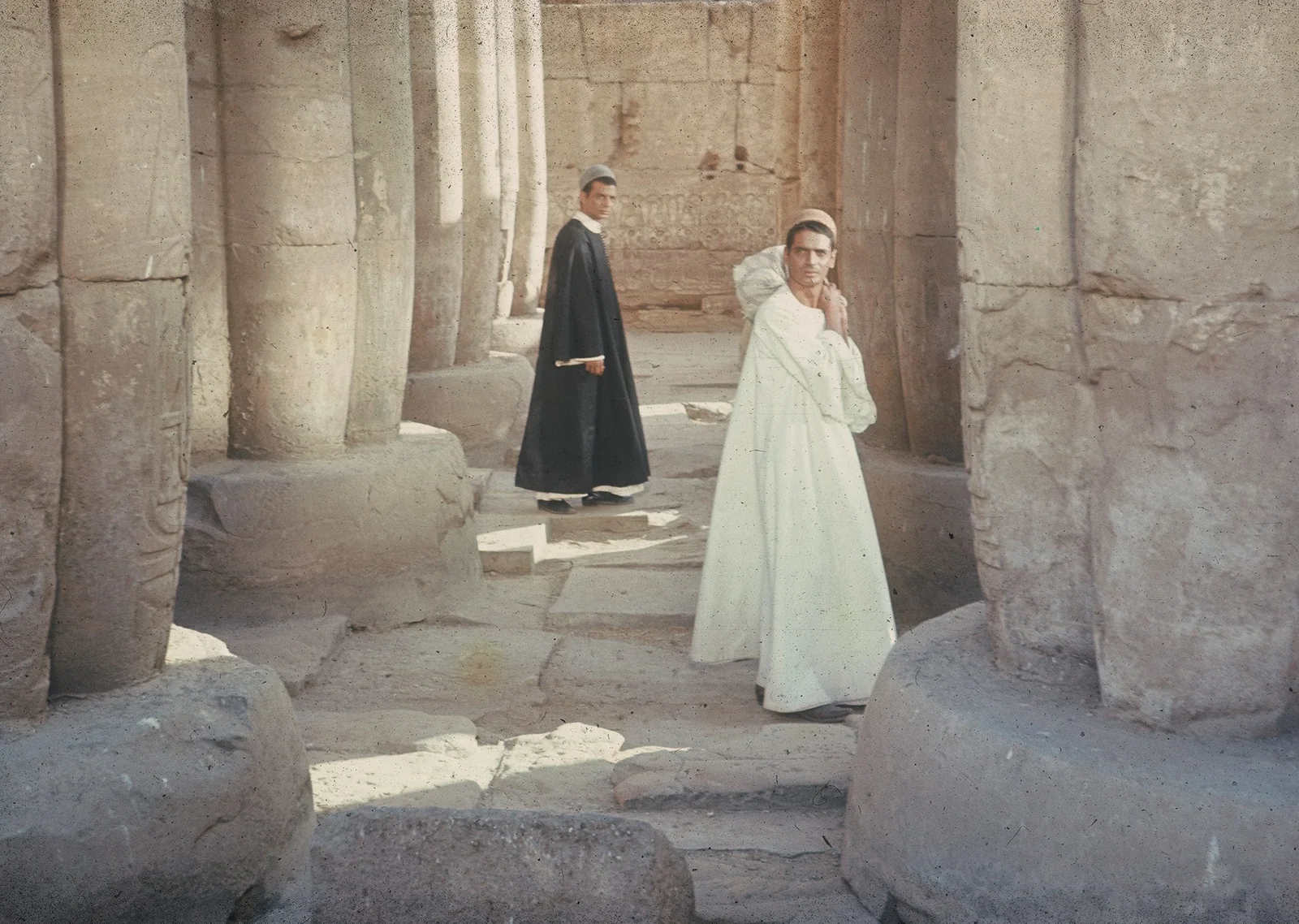 Two men in black and white robes stand among massive weathered stone columns in The Night of Counting the Years.