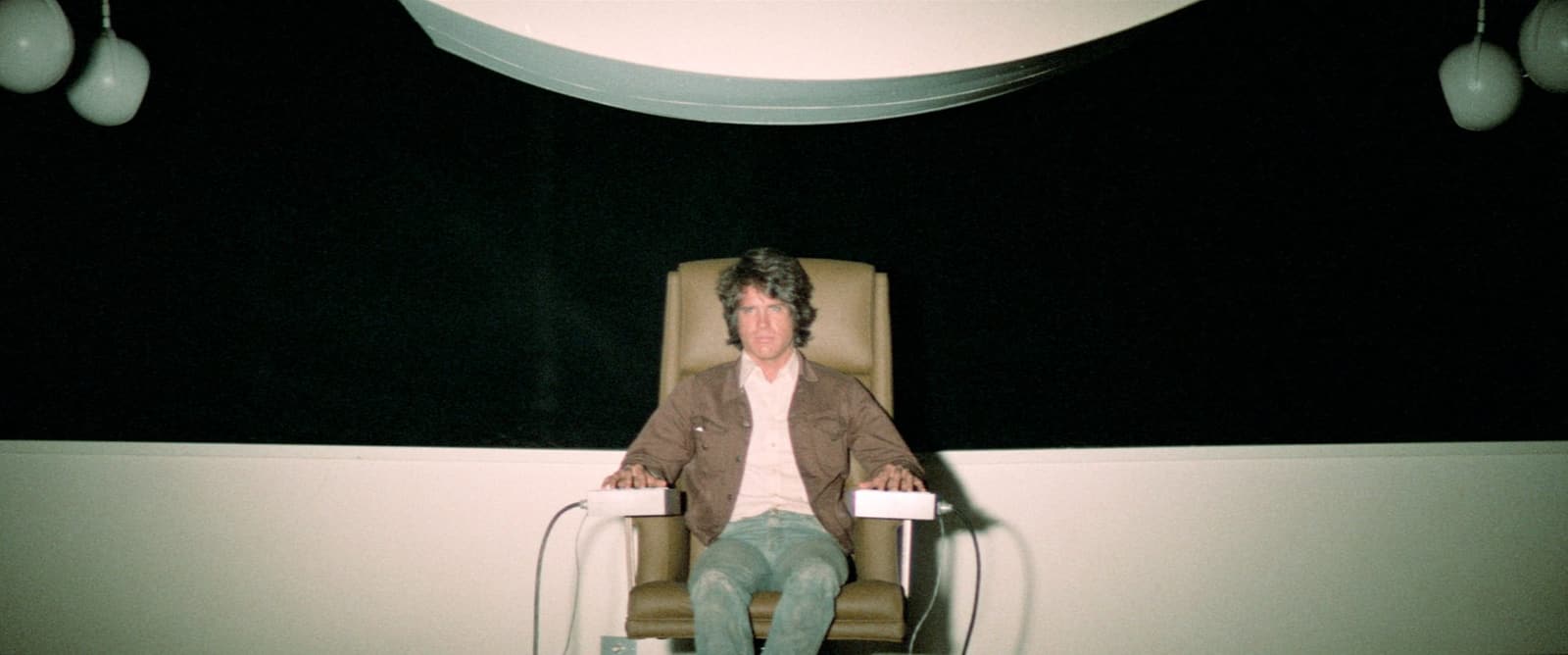 Warren Beatty as Joe Frady sits alone in a stark testing chair inside a dark screening room in The Parallax View.
