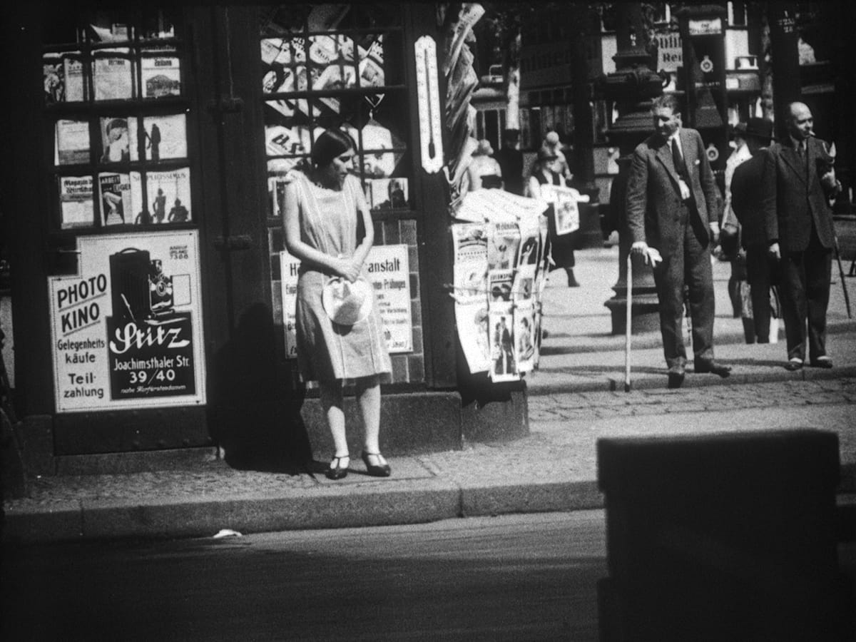 A young woman in a light summer dress waits at a Berlin shopfront beside a large Photo Kino camera advertisement in People on Sunday.