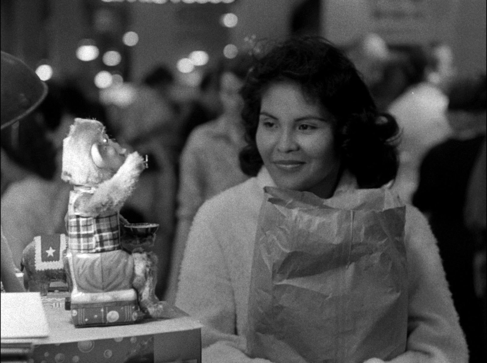 Yvonne Williams smiles at a bubble-blowing toy monkey beside a market display in the black-and-white film The Exiles.