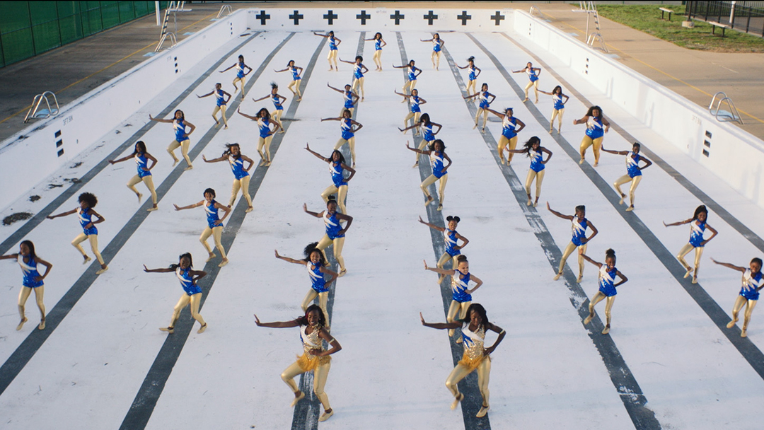 The Lionesses drill team, wearing blue and gold uniforms, performs in rows across a drained outdoor swimming pool in The Fits.